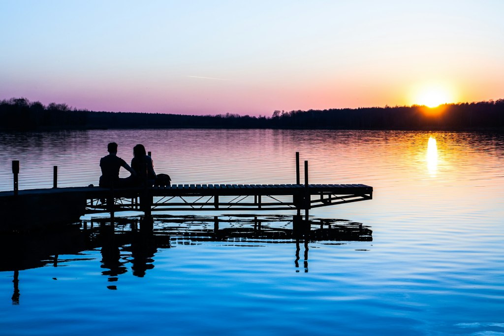 Silhouettes of two people sitting side by side on a dock at dusk