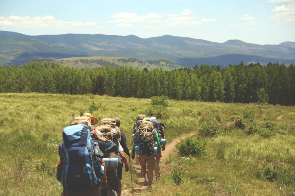 A group of young hikers with backpacks walking a dirt trail through a green meadow
